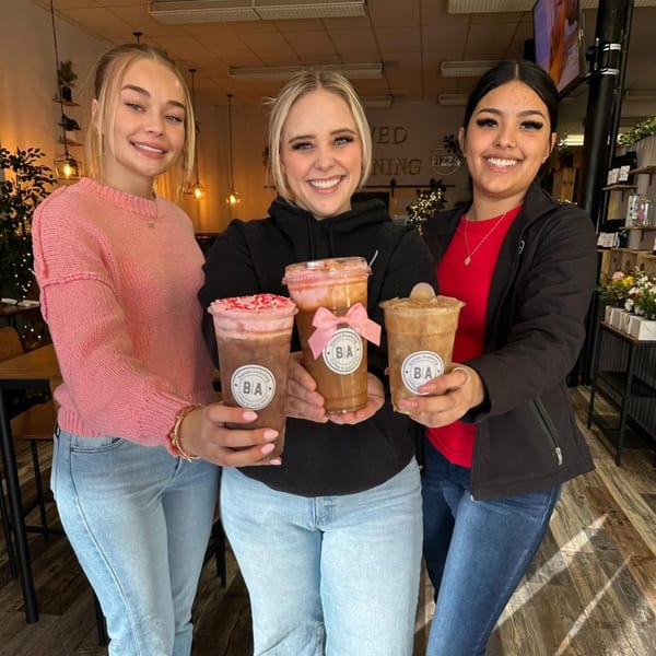 Three smiling women holding Brewed Awakening coffee drinks with various toppings in a cafe.