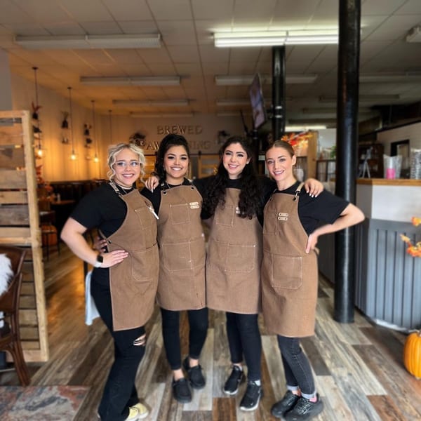 Four smiling baristas in brown aprons stand together inside Brewed Awakening Coffee and Bake Shop.