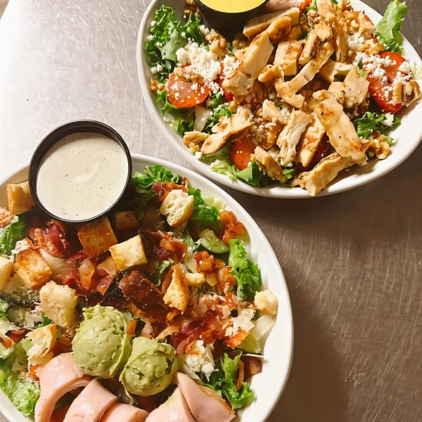 Two vibrant salads on a metal surface, one with chicken, the other with avocado and ham.