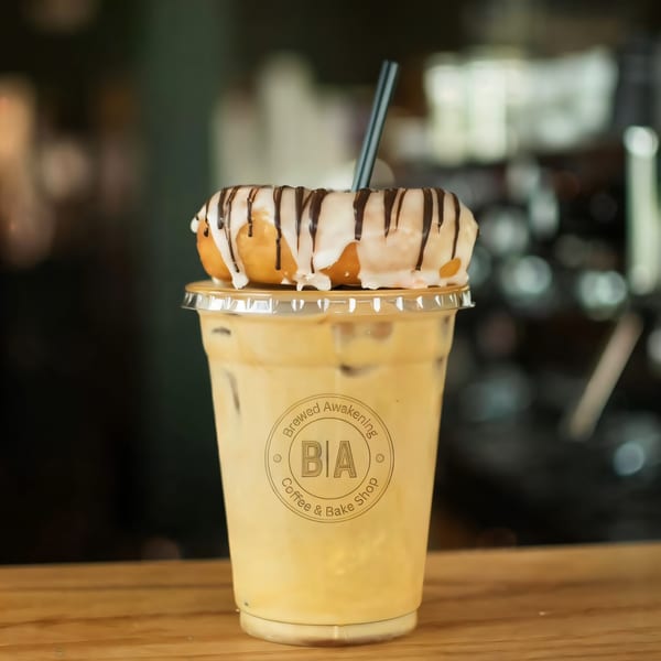 Iced coffee with a Brewed Awakening logo, topped with a glazed donut drizzled with chocolate.
