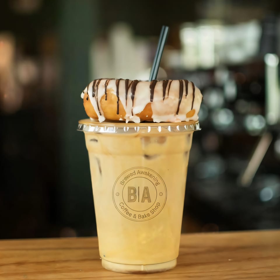 Iced coffee with a Brewed Awakening logo, topped with a glazed donut drizzled with chocolate.