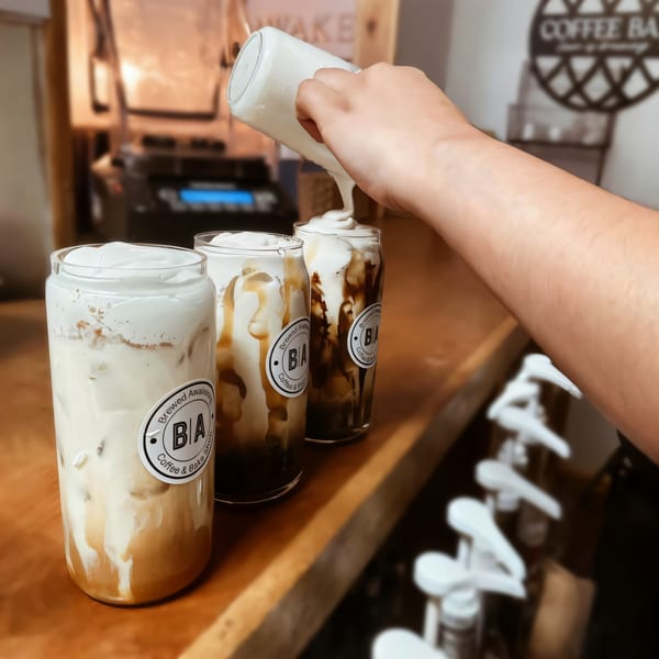 Hand pouring whipped cream onto three iced coffee drinks with Brewed Awakening labels on a counter.