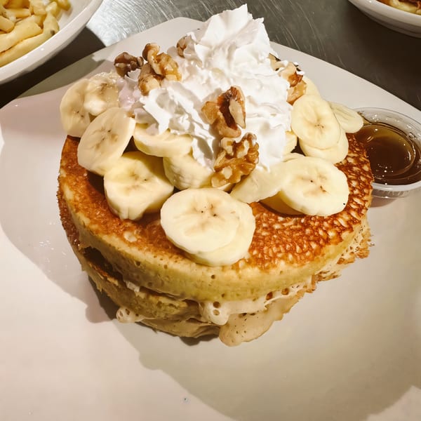 Stack of pancakes topped with whipped cream, sliced bananas, walnuts, and a side of syrup.
