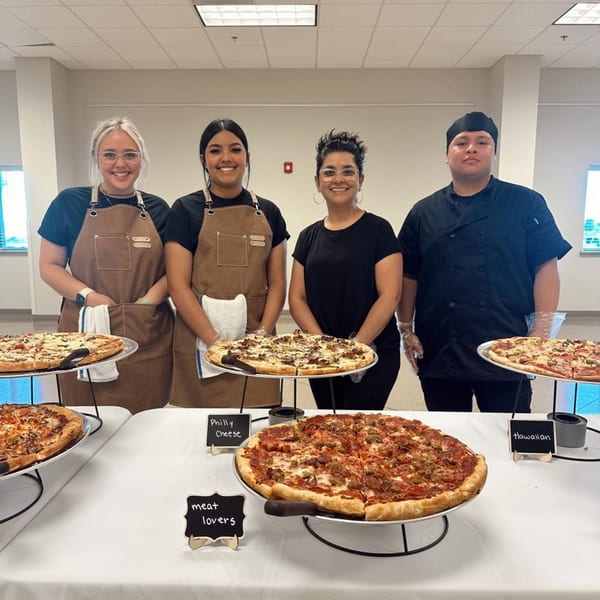Four smiling staff members stand behind a table laden with various pizzas, including meat lovers and Hawaiian.